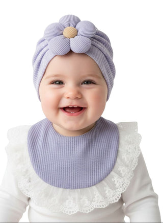 Baby wearing a purple knit hat with a flower on top and matching bib, smiling against a white background.