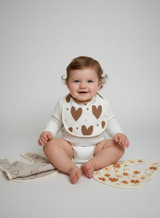Baby sitting on a white surface wearing a heart-patterned bib with additional bibs and a blanket around.