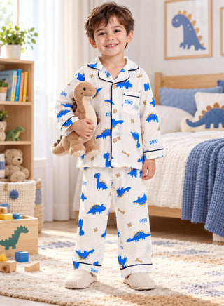 Boy wearing a white full-sleeve dinosaur print pajama set with bright blue dinosaur patterns, navy piping, button-down shirt and matching full-length pants.