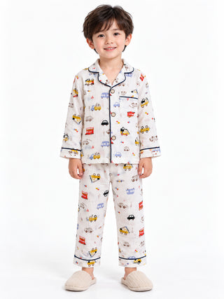 Boy wearing a white multicolor vehicle print pajama set featuring cars, buses and trucks, navy blue piping, a button-down shirt and matching full-length pants.