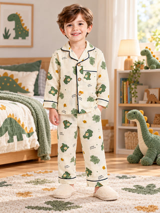 Boy wearing a cream pajama set with cute green dinosaur prints, Little Dino lettering, navy blue piping, full-sleeve button-down shirt and matching pants.