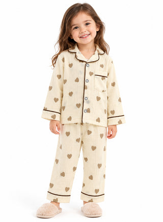 Girl wearing a cream pajama set with small brown heart prints, dark brown contrast piping, full-sleeve button-down shirt and matching pants.
