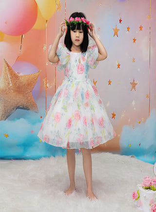A young girl stands barefoot on a white faux-fur rug wearing a white tulle dress with pink, blue, and green floral print and puff sleeves. She is posed in front of a whimsical backdrop featuring balloons, stars, clouds, and a glittery star.