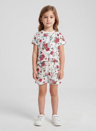 A young girl stands against a light gray studio background wearing a short-sleeve white romper with a red and blue floral print, a matching drawstring waist, and white sneakers.