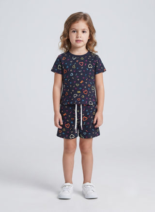 A young girl stands against a light gray studio background wearing a matching short-sleeve t-shirt and shorts set in dark navy blue with a colorful heart pattern, paired with white sneakers.